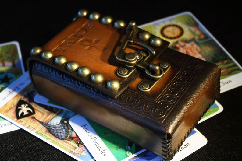 May include: A brown leather tarot card deck box with a brass clasp and decorative studs. The box is closed and has the word "Tarot" embossed on the front.