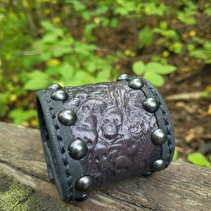 May include: A black leather bracelet with a textured design and silver studs. The bracelet is on a wooden surface.