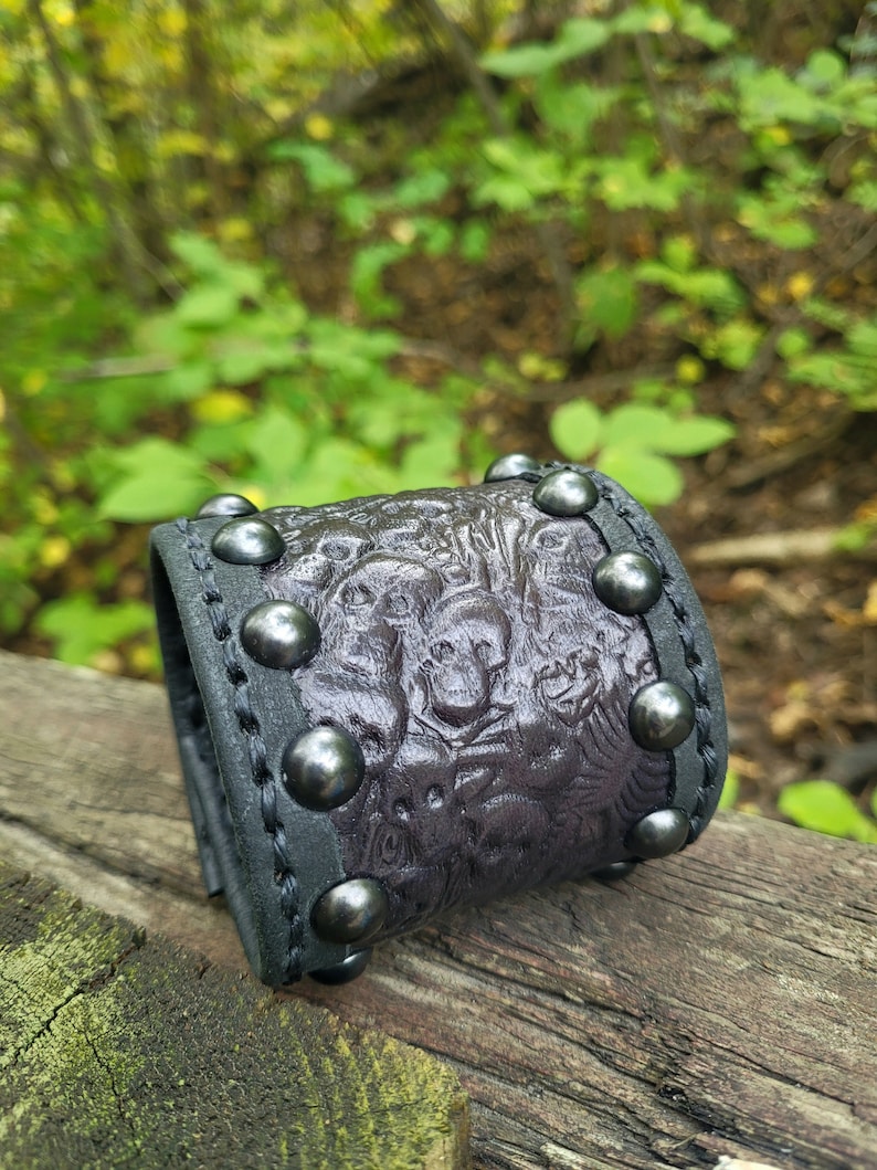 May include: A black leather bracelet with a textured design and silver studs. The bracelet is on a wooden surface.