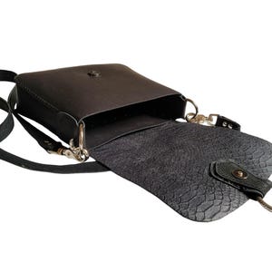 May include: A black leather crossbody bag with a flap closure and a silver buckle. The bag has a textured lining and a silver ring on the strap.