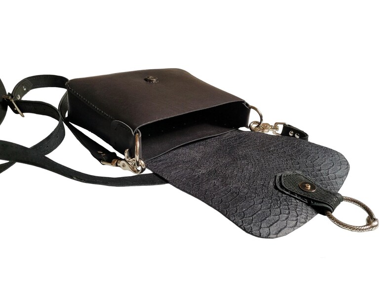 May include: A black leather crossbody bag with a flap closure and a silver buckle. The bag has a textured lining and a silver ring on the strap.
