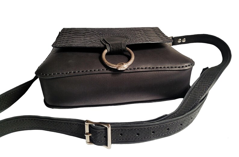 May include: Black leather crossbody bag with a snake-shaped ring detail and an adjustable strap.