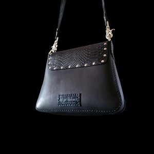 May include: Black leather shoulder bag with a snakeskin pattern flap and silver studs. The bag has a silver buckle closure and a brand label that reads "Mystic Maverick Leather & Goods".