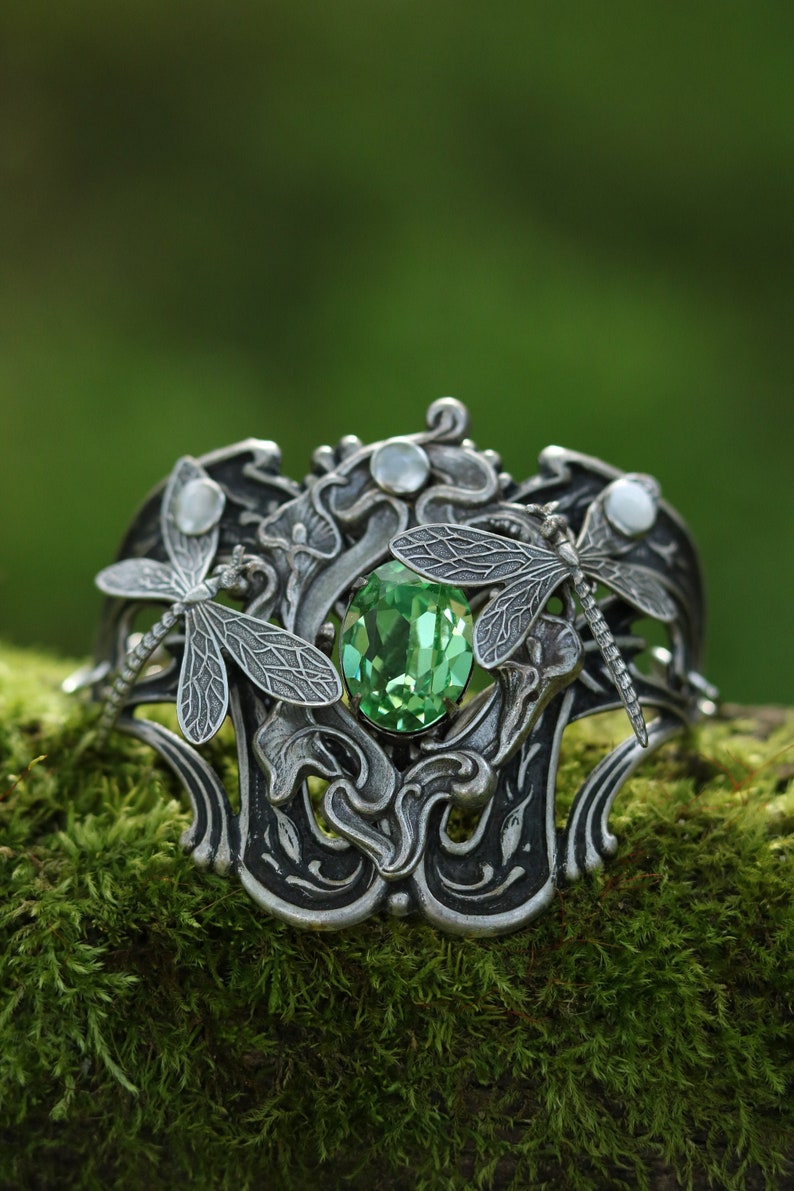 May include: A silver bracelet featuring a green gemstone and two dragonfly accents. The bracelet is resting on a bed of green moss.