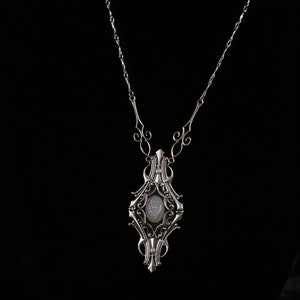 May include: Silver-toned pendant necklace with an ornate, Art Nouveau-inspired design. The pendant features a central oval-shaped stone with a shimmering, iridescent appearance. The necklace has a delicate chain and decorative flourishes.