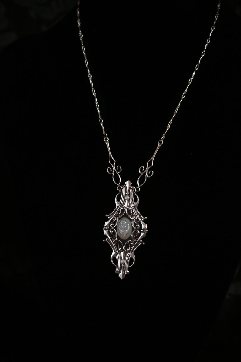 May include: Silver-toned pendant necklace with an ornate, Art Nouveau-inspired design. The pendant features a central oval-shaped stone with a shimmering, iridescent appearance. The necklace has a delicate chain and decorative flourishes.