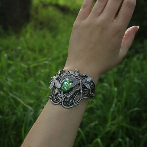 May include: A silver bracelet featuring a green gemstone and dragonfly details. The bracelet is worn on a person's wrist against a backdrop of green grass.