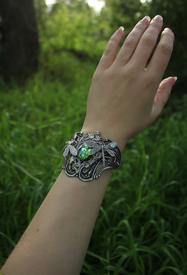 May include: A silver bracelet featuring a green gemstone and dragonfly details. The bracelet is worn on a person's wrist against a backdrop of green grass.