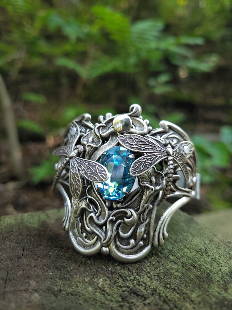 May include: A silver bracelet featuring a blue gemstone and dragonfly details. The bracelet is intricately designed with swirling patterns and delicate wings.