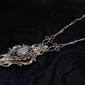 May include: A silver-toned necklace with an ornate pendant featuring a central, iridescent stone. The pendant has an elaborate, filigree design with swirling patterns and decorative elements. The necklace chain is delicate and complements the pendant's intricate details.