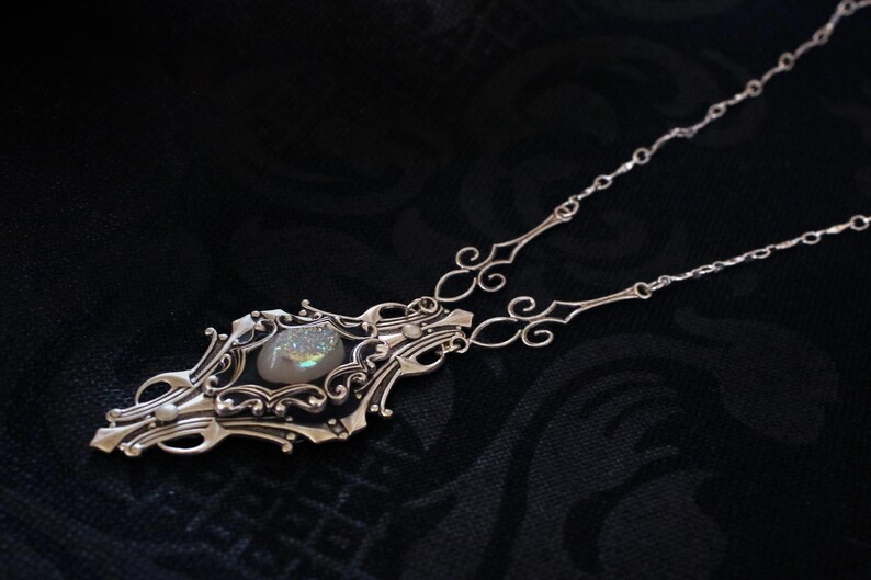 May include: A silver-toned necklace with an ornate pendant featuring a central, iridescent stone. The pendant has an elaborate, filigree design with swirling patterns and decorative elements. The necklace chain is delicate and complements the pendant's intricate details.
