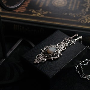 May include: A silver pendant necklace with an ornate, filigree design. The pendant features a central, oval-shaped, light-colored stone. The necklace is displayed on a black box, with a dark background and a book visible in the background.