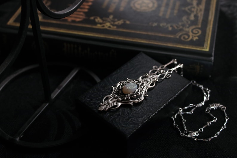 May include: A silver pendant necklace with an ornate, filigree design. The pendant features a central, oval-shaped, light-colored stone. The necklace is displayed on a black box, with a dark background and a book visible in the background.