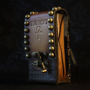 May include: A brown leather tarot card deck box with brass accents and an ankh symbol. The box has the word "Tarot" embossed on the front.