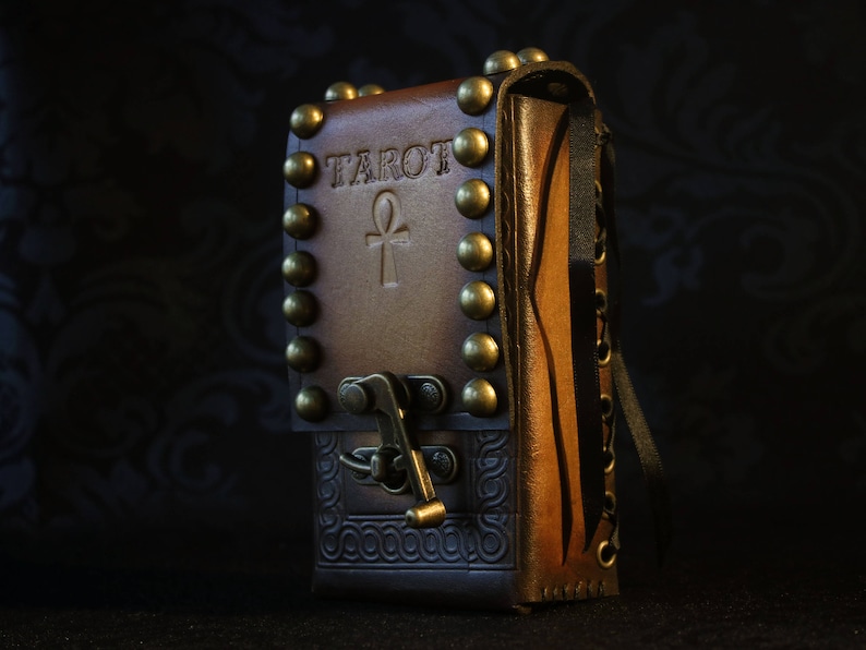 May include: A brown leather tarot card deck box with brass accents and an ankh symbol. The box has the word "Tarot" embossed on the front.