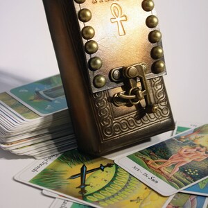 May include: A brown leather tarot card deck box with a brass clasp and metal studs. The box is embossed with the word "TAROT" and an ankh symbol. The box is sitting on top of a stack of tarot cards.