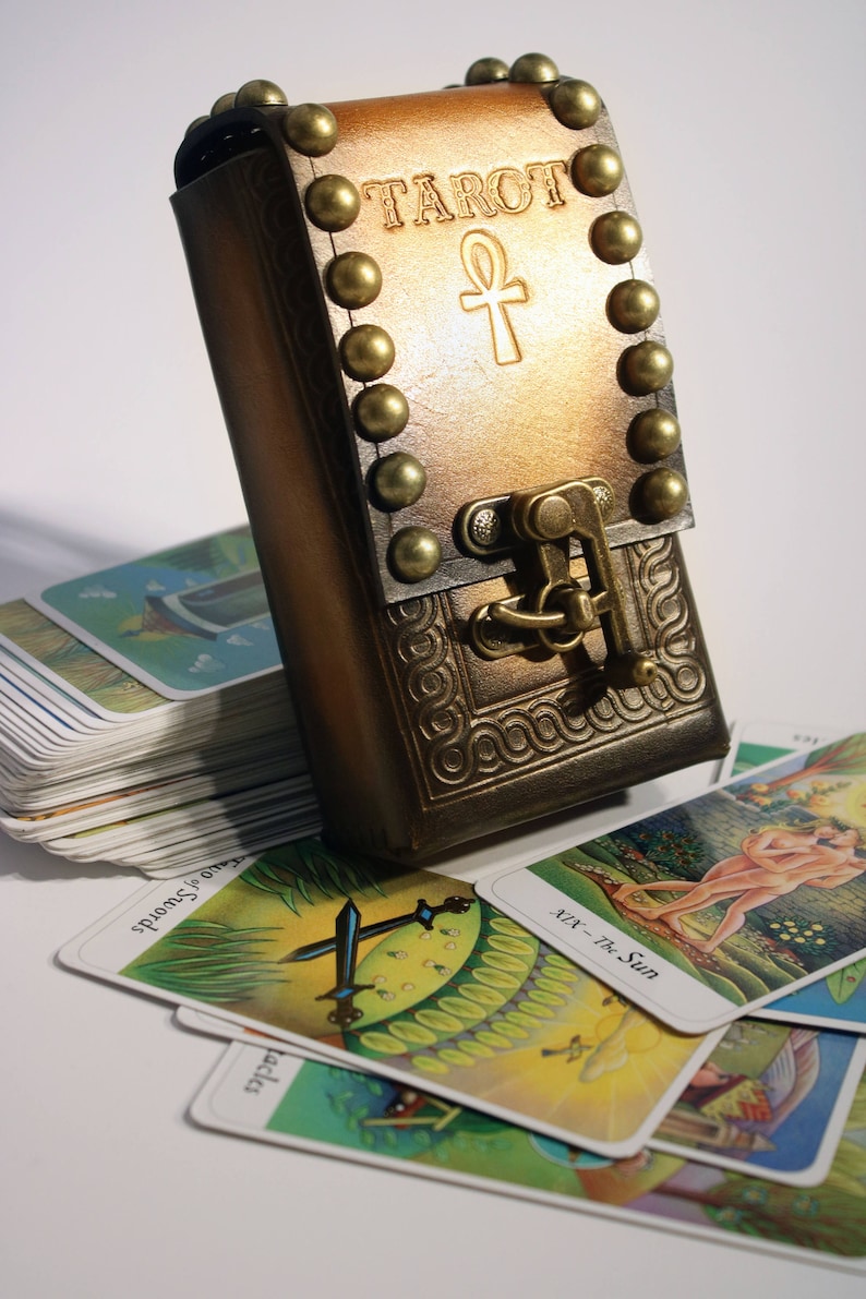 May include: A brown leather tarot card deck box with a brass clasp and metal studs. The box is embossed with the word "TAROT" and an ankh symbol. The box is sitting on top of a stack of tarot cards.