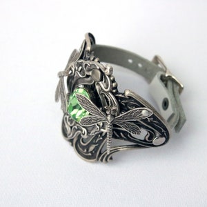 May include: A silver bracelet featuring a dragonfly design and a green gemstone. The bracelet is made of leather and metal.