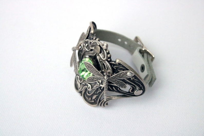 May include: A silver bracelet featuring a dragonfly design and a green gemstone. The bracelet is made of leather and metal.