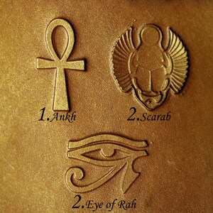 May include: Close-up of three gold-colored metal stamps with Egyptian symbols. The stamps are labeled "1. Ankh", "2. Scarab", and "2. Eye of Rah".
