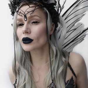 May include: A woman with long, silver hair wears a black crown with black roses and a black and silver chain necklace. She has black wings and dark makeup.
