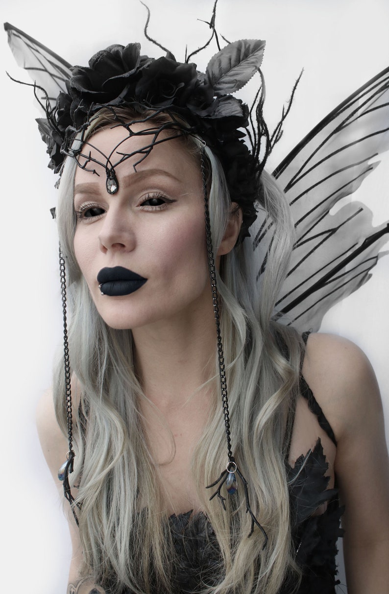 May include: A woman with long, silver hair wears a black crown with black roses and a black and silver chain necklace. She has black wings and dark makeup.