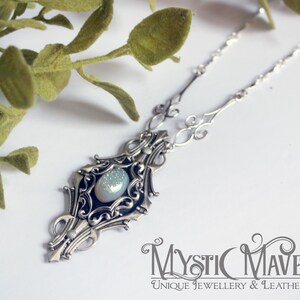 May include: A silver-toned necklace with an ornate pendant featuring a central, iridescent stone. The pendant is surrounded by intricate filigree detailing. The necklace is displayed on a white surface with green foliage in the background. The Mystic Maverick logo is visible.
