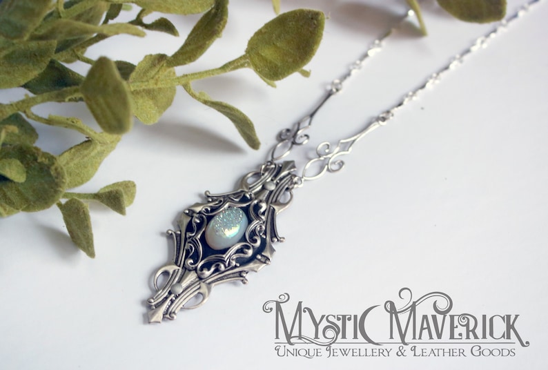 May include: A silver-toned necklace with an ornate pendant featuring a central, iridescent stone. The pendant is surrounded by intricate filigree detailing. The necklace is displayed on a white surface with green foliage in the background. The Mystic Maverick logo is visible.
