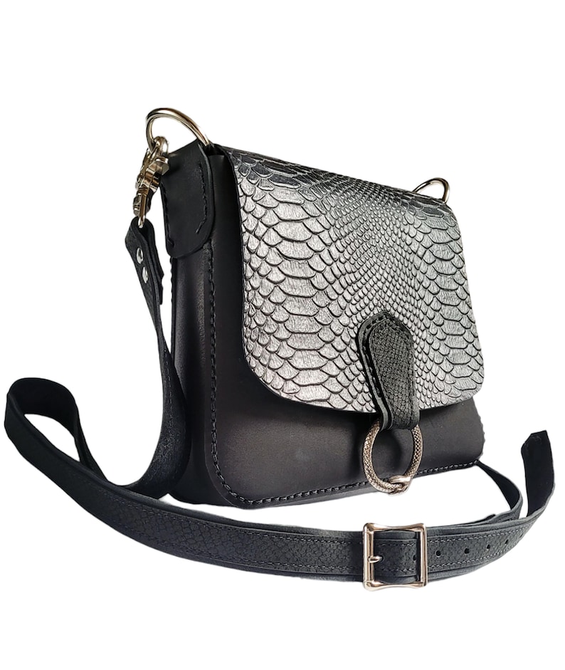 May include: A black leather crossbody bag with a silver snakeskin flap and a gold ring closure. The bag has a long adjustable strap.