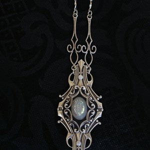 May include: Silver-toned pendant necklace with an ornate, Art Nouveau-inspired design. The pendant features a central oval-shaped stone with a shimmering, iridescent surface, surrounded by intricate filigree and decorative elements. The necklace hangs on a delicate chain.