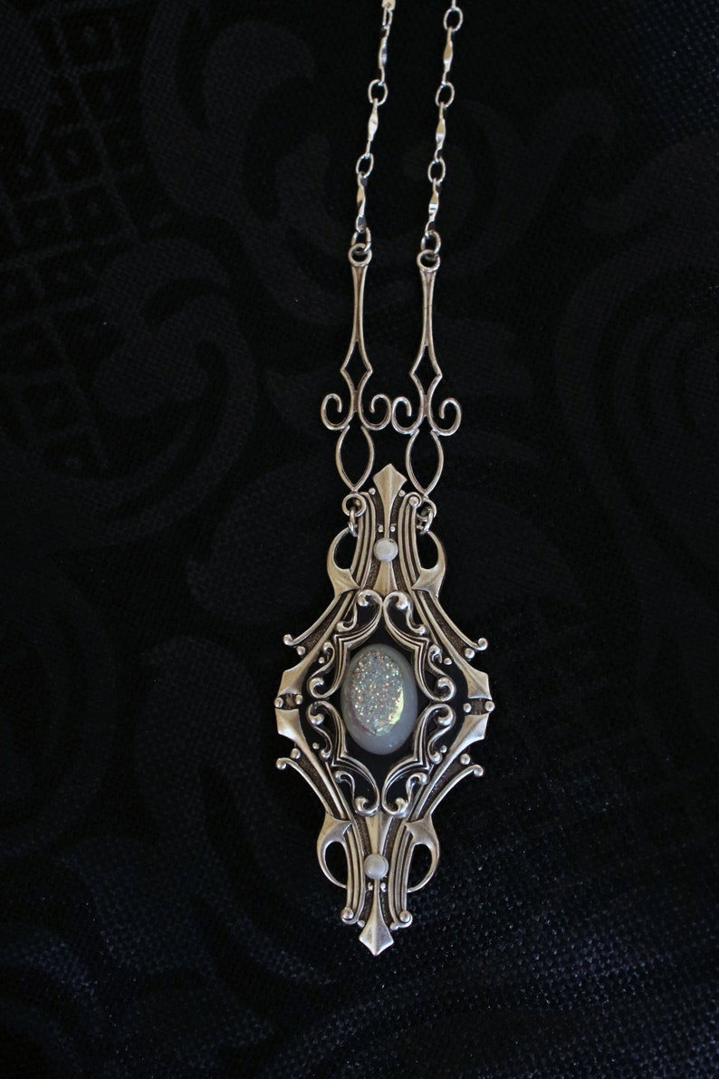 May include: Silver-toned pendant necklace with an ornate, Art Nouveau-inspired design. The pendant features a central oval-shaped stone with a shimmering, iridescent surface, surrounded by intricate filigree and decorative elements. The necklace hangs on a delicate chain.