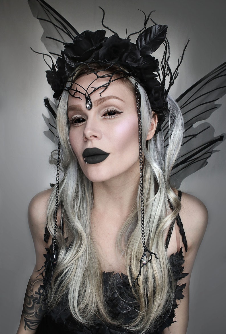May include: A woman with long, silver hair wears a black floral crown with a black crystal pendant. She has black eyeliner and dark lipstick. She is wearing a black dress with a lace overlay and black wings.