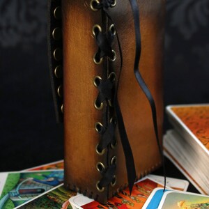 May include: A brown leather tarot card deck box with a black ribbon and brass eyelets. The box is laced up the side with a black ribbon. The box is sitting on top of a stack of tarot cards.