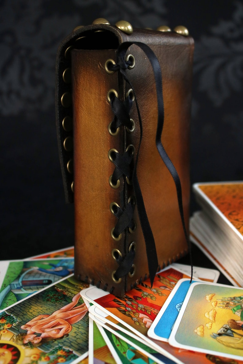 May include: A brown leather tarot card deck box with a black ribbon and brass eyelets. The box is laced up the side with a black ribbon. The box is sitting on top of a stack of tarot cards.
