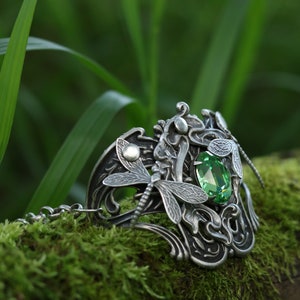 May include: A silver bracelet featuring a dragonfly design and a green gemstone. The bracelet is resting on a bed of moss.