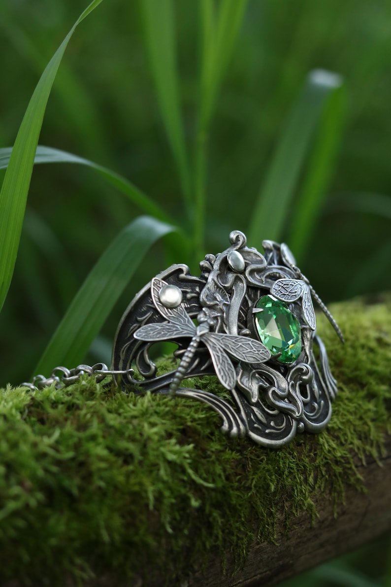May include: A silver bracelet featuring a dragonfly design and a green gemstone. The bracelet is resting on a bed of moss.