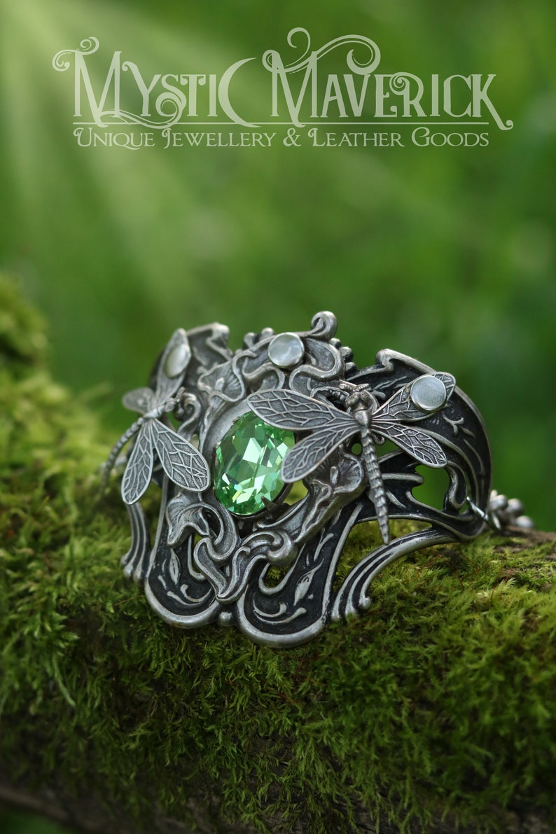 May include: A silver bracelet featuring a green gemstone and dragonfly accents. The bracelet is intricately designed with floral motifs and has a delicate chain.