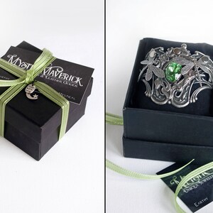 May include: A green gemstone ring with dragonfly accents, set in a silver band. The ring is displayed in a black gift box with a green ribbon and a tag that reads "Mystical Maverick & Leather Goods".