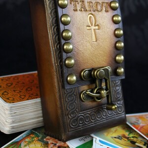 May include: A brown leather tarot card deck box with a brass clasp and studs. The box has the word "TAROT" and an ankh symbol embossed on the front. The box is open and a deck of tarot cards is visible.