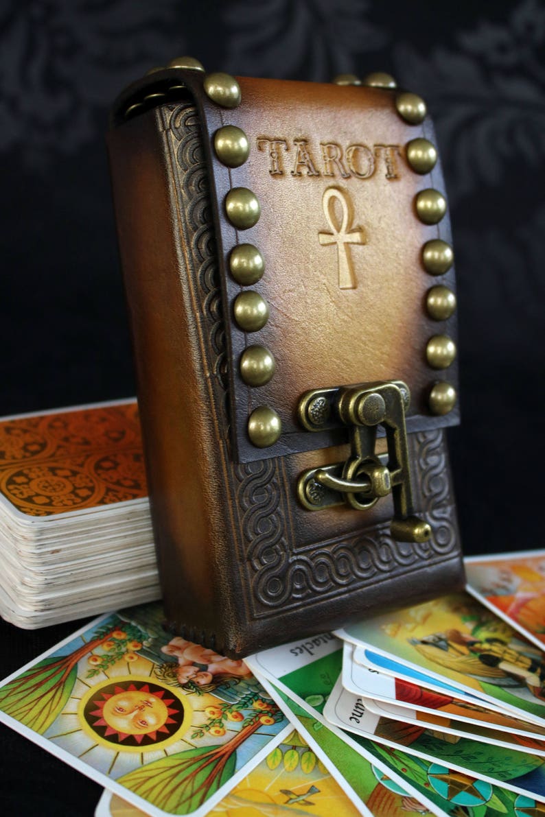 May include: A brown leather tarot card deck box with a brass clasp and studs. The box has the word "TAROT" and an ankh symbol embossed on the front. The box is open and a deck of tarot cards is visible.