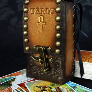 May include: A brown leather tarot card deck box with brass studs and a clasp. The box has the word "TAROT" and an ankh symbol embossed on the front. The box is sitting on top of a stack of tarot cards.