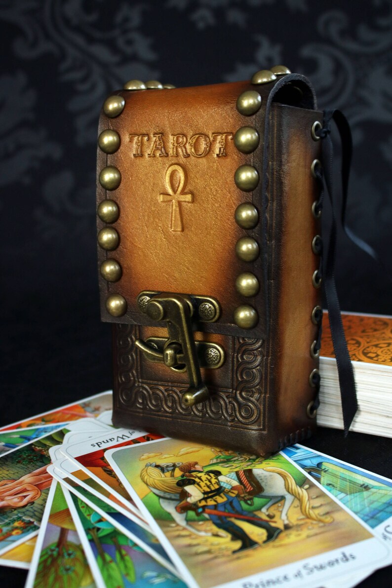 May include: A brown leather tarot card deck box with brass studs and a clasp. The box has the word "TAROT" and an ankh symbol embossed on the front. The box is sitting on top of a stack of tarot cards.