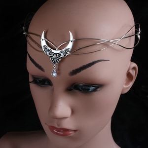 May include: A silver wire headband with a crescent moon design and a dangling crystal. The headband is worn on a mannequin's head.