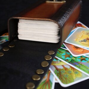 May include: A brown leather box with a brass clasp, open to reveal a stack of cards with colorful illustrations. The box is decorated with a pattern of small circles and has a black leather strap with brass studs.