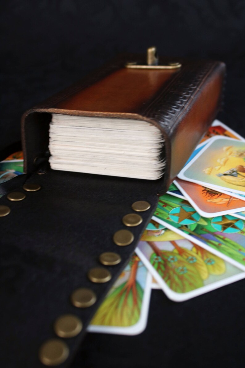 May include: A brown leather box with a brass clasp, open to reveal a stack of cards with colorful illustrations. The box is decorated with a pattern of small circles and has a black leather strap with brass studs.