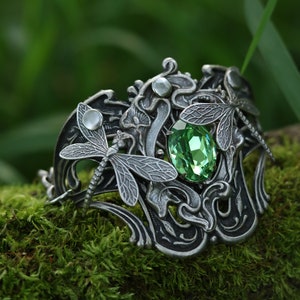 May include: A silver bracelet featuring a green gemstone and dragonfly details. The bracelet is resting on a bed of green moss.