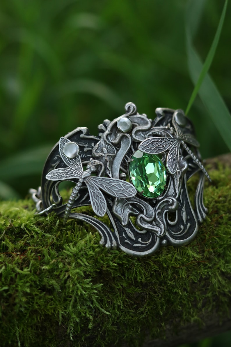 May include: A silver bracelet featuring a green gemstone and dragonfly details. The bracelet is resting on a bed of green moss.