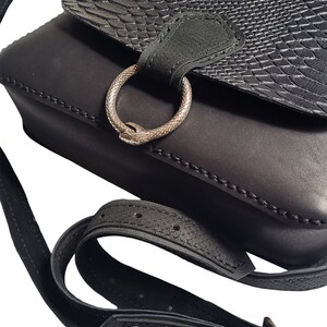 May include: Black leather crossbody bag with a snake-embossed flap and a silver ring closure. The bag has a long adjustable strap with a buckle.