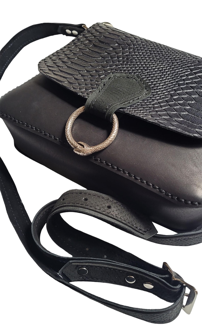 May include: Black leather crossbody bag with a snake-embossed flap and a silver ring closure. The bag has a long adjustable strap with a buckle.