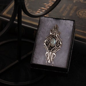 May include: Ornate silver pendant with a central iridescent stone, displayed in a black gift box. The pendant features intricate filigree detailing and a delicate chain. The background includes a dark book, suggesting a mystical or gothic theme.